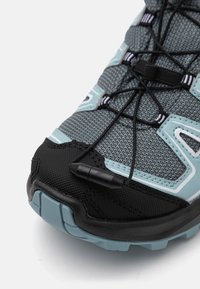 Grey athletic shoe with a mesh upper, black rubber toe cap, light blue accents, and a lace-lock system for secure fit.