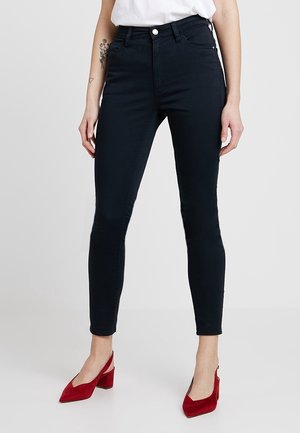 Kvinde iført sorte skinny jeans, hvid top og røde hælede sko, står med hænder afslappet ned langs siden mod en ensfarvet baggrund.