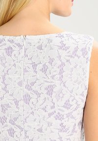 Tissu en dentelle blanche à motifs floraux superposés sur une base lavande, doté d'un décolleté carré et d'une fermeture éclair au dos.