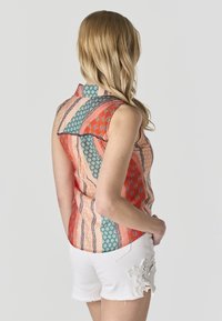Mujer con cabello rubio, vestida con una blusa sin mangas de estampado multicolor y pantalones cortos blancos con detalles de encaje, de pie contra un fondo claro liso.