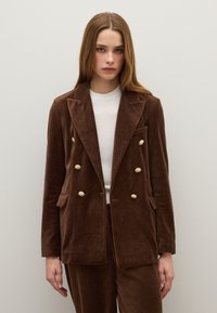 Bruine fluwelen dubbelbreasted blazer met gouden knopen, ingesneden revers, twee voorzakken en een iets oversized pasvorm. Witte binnenlaag zichtbaar.