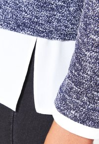 Stickad topp med ett strukturerat mönster i mörkblått och vitt, med ett vitt undertröja som är synligt vid fållen och ärmarna.