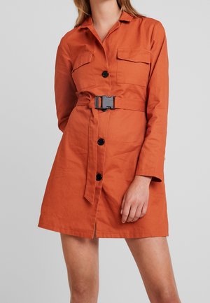 Mujer con un vestido naranja con botones, dos bolsillos en el pecho y un cinturón con hebilla de plástico negro, sobre un fondo gris liso.