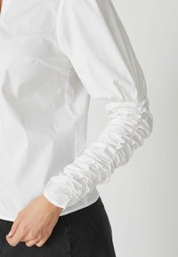 Blusa bianca con colletto, maniche lunghe con dettagli arricciati e un tessuto leggero e liscio, indossata con abbigliamento scuro.