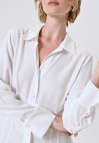 Witte, knoopsluiting blouse met een zachte textuur, lange mouwen en een klassieke kraag. Heeft een subtiele glans en een ontspannen pasvorm.