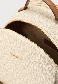 Sac à dos en toile beige avec un motif texturé de lettres entrelacées, accents en cuir marron, et fermetures éclair dorées. Nom de la marque visible.