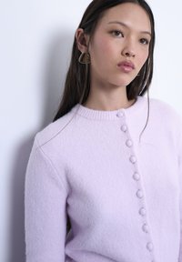 Νεαρή γυναίκα με μακριά σκούρα μαλλιά, που φοράει γεωμετρικά σκουλαρίκια και ένα ανοιχτό μοβ πουλόβερ με κουμπιά, κοιτάζοντας ελαφρώς προς τα πλάγια.