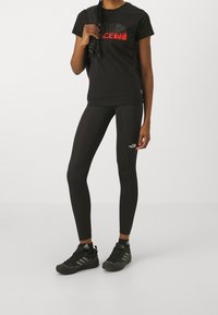 Czarna koszulka z czerwoną grafiką logo, czarne legginsy i czarne buty sportowe. Gładki materiał, dopasowany krój i lekkie materiały.