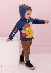 Batole chlapec v modré mikině s dinosaurem s barevnými hřbety a hnědých kalhotách, jak vyvažuje jednou rukou na bílém kvádru.