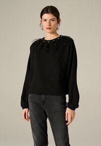 Felpa nera con un accento in piuma testurizzata sul colletto, con maniche lunghe e un fit rilassato. Abbinata a jeans scuri.