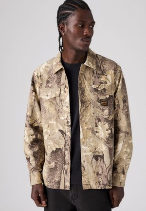 Homme aux cheveux tressés portant une chemise beige et marron à motifs boutonnée par-dessus un t-shirt noir, regardant vers la gauche sur un fond uni.
