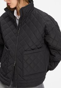 Giacca nera trapuntata con colletto alto, zip frontale e grandi tasche a toppa. Presenta un motivo a diamante testurizzato e un tessuto morbido.