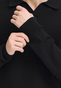 Čierny pletený pulóver s výstrihom do V a golierom. Detailný záber na ruky upravujúce rukáv, zobrazuje rebrované manžety a hladkú textúru.