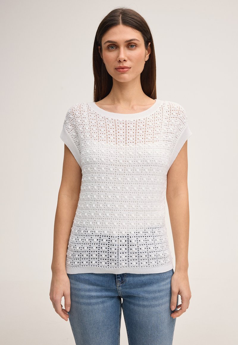 Femme aux cheveux bruns raides portant un haut en tricot blanc à manches courtes avec des motifs ajourés et un jean bleu, debout devant un fond uni.