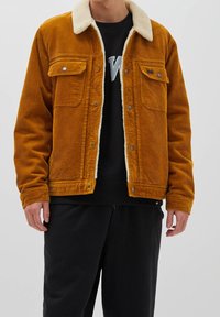 Giacca in velluto a coste color ambra con collo in shearling, tasche frontali a patta e bottoni a pressione. Indossata sopra un felpa nera e pantaloni.