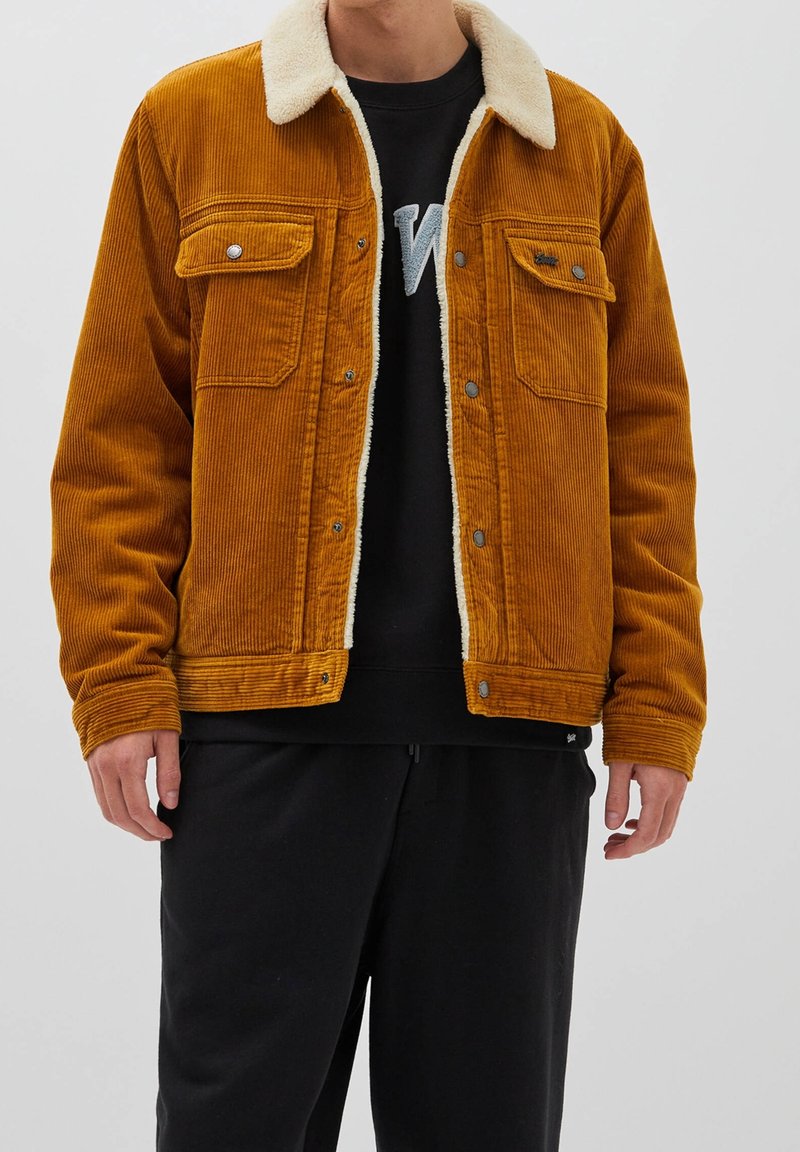 Giacca in velluto a coste color ambra con collo in shearling, tasche frontali a patta e bottoni a pressione. Indossata sopra un felpa nera e pantaloni.