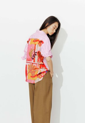 Femme aux longs cheveux bruns portant une chemise rose ample avec des motifs floraux abstraits colorés dans le dos et un pantalon marron à jambes larges, debout de profil.