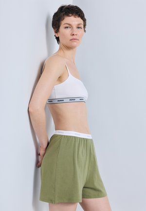 Femeie cu păr scurt, purtând un sutien sport alb cu marcă și pantaloni scurți verde măsliniu, stând în partea laterală, cu fața spre perete gri deschis.