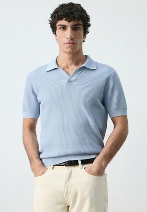 Joven con cabello rizado, vestido con una camiseta tipo polo de punto de manga corta azul claro y pantalones color crema, con las manos en los bolsillos, frente a un fondo liso.