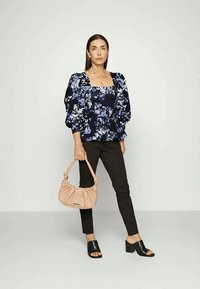 Blusa floral azul-marinho com mangas bufantes, calças pretas ajustadas e uma mala bege. Slides de salto aberto pretos.