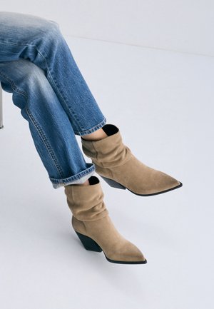 Personne portant un jean bleu et des bottines beige en daim avec des bouts pointus et des talons épais, assise sur une chaise devant un fond uni.