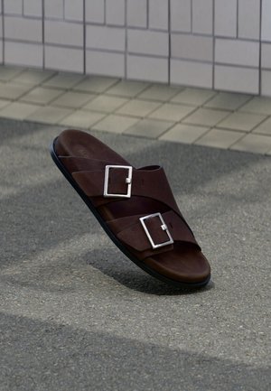 Rjava usnjena sandal z dvema srebrnima zaponkama, ki počiva na teksturirani sivi tlakovci blizu bež obložene stene.