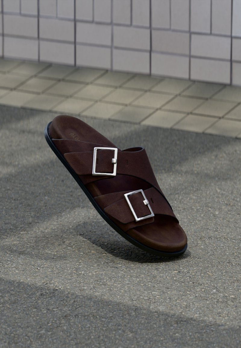 Sandale en cuir marron avec deux boucles argentées reposant sur un pavement gris texturé près d'un mur carrelé beige.