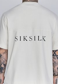 Lühikeste varrukatega, kreemjas valges T-särk, mille tagasi disainil on lihtne "SIKSILK" julgete mustade tähtedega. Valmistatud pehmest ja kergest materjalist.
