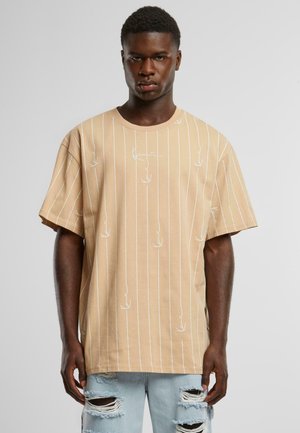Tan oversized T-shirt with white vertical stripes and small leaf motifs, made of soft cotton fabric, featuring a round neckline and short sleeves.