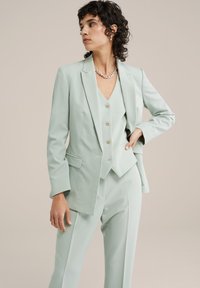 Mint zelena tailored obleka, ki vključuje strukturirano blazer, prilegajoč brezrokavnik s gumbi in hlače ravnega kroja z nagibom spredaj.