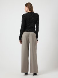 Donna in piedi di spalle, che indossa un top nero a maniche lunghe testurizzato e pantaloni beige a vita alta e gamba larga, con tacchi neri.