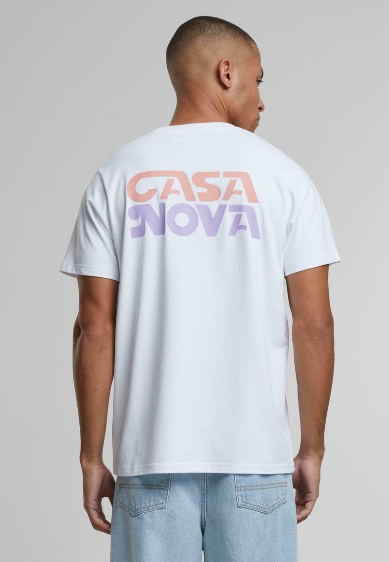 Balta kokvilnas T-kreklu ar izteiksmīgu grafisko apdruku aizmugurē, kurā ir teksts "CASA NOVA" korallzilos un purpura toņos. Īsās piedurknes, brīva piegriezuma.