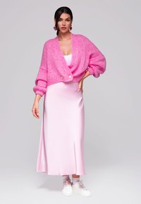 Růžový pletený zkrácený cardigan s objemovými rukávy, kombinovaný s dlouhou růžovou saténovou šaty a bílými teniskami, na neutrálním pozadí.