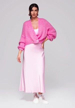 Rosa gestrickte kurze Strickjacke mit übergroßen Ärmeln, kombiniert mit einem langen rosa Satin-Kleid und weißen Sneakers, vor einem neutralen Hintergrund.