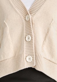 Beige cardigan made of soft knit fabric, featuring a deep V-neck, ribbed cuffs, and three large white buttons at the front.