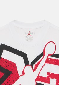 Λευκό βαμβακερό T-shirt με μαύρα και κόκκινα γραφικά, που απεικονίζουν τη σιλουέτα ενός παίκτη που πηδάει. Ορατό τυπωμένο λογότυπο και ετικέτα φροντίδας.