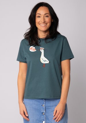 Teal korte mouwen t-shirt met een gestileerde witte vogelafbeelding en een tekstballon met de tekst "MOIN!" De print is voorzien van kleurrijke accenten.