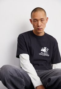 Dunkelgraues T-Shirt mit weißem grafischem Druck, übereinander getragen mit einem weißen Langarmshirt. Lässige Passform mit Kurzärmeln, kombiniert mit lockeren grauen Jeans.