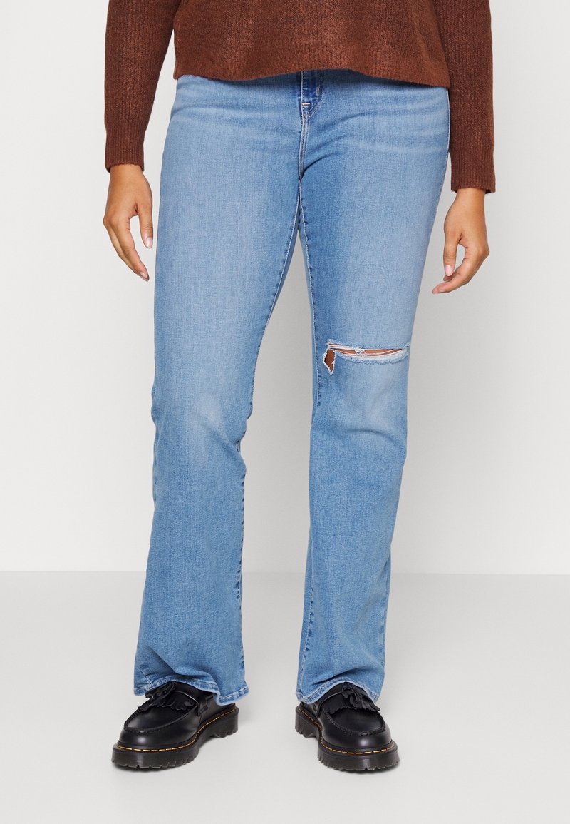 Personne portant un jean bleu clair avec un déchirure au genou, un pull marron et des chaussures noires épaisses, debout devant un fond blanc uni.