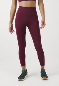 Bordó, magas derekú leggings rugalmas anyagból, lapos varrásokkal, zöld sportcipőkkel párosítva, amelynek textúrázott talpa van.