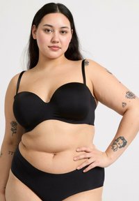 Soutien-gorge noir rembourré avec des bretelles fines réglables et une texture lisse. Il présente un design structuré pour un bon maintien. Le modèle porte des bas de bikini noirs assortis.