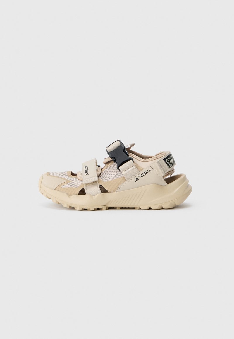 Sandalias de exterior beige con parte superior de malla, correas ajustables y suela de goma texturizada para mayor agarre. Presenta un acento negro en el talón.