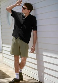 Black textured short-sleeve button-up shirt with black buttons, paired with olive green shorts and black shoes, on wooden deck against white wall.