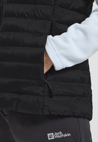 Gilet imbottito nero con tasca con zip, caratterizzato da una texture liscia. Sotto, una manica in pile azzurro chiaro è parzialmente visibile.