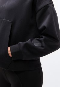 Άτομο που φοράει μαύρο φούτερ με το αριστερό του χέρι στην μπροστινή τσέπη, εμφανίζοντας ριμπ μανσέτες και διακριτικές αντανακλαστικές ρίγες στο μανίκι.