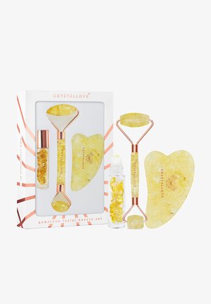 Zestaw kosmetyków do twarzy z kamieniami szlachetnymi, zawierający żółte narzędzia z różowymi złotymi akcentami, wałek, gua sha oraz szklany pojemnik na serum, w eleganckim opakowaniu.