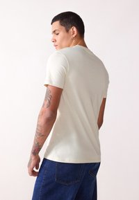 Light beige fitted t-shirt with short sleeves, round neckline, soft texture. Model wears dark blue denim shorts, showcasing a tattoo on the arm.