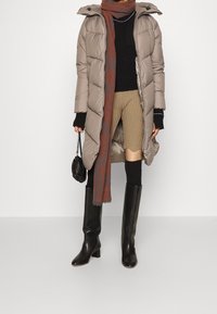 Person iført lang beige pufferjakke, sort sweater, tan rillede nederdel, sorte knæhøje støvler, tørklæde, holder en lille sort håndtaske.