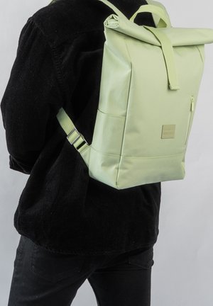 Personne portant un sac à dos à enroulement vert clair avec une poche avant zippée, vêtue d'une veste en velours côtelé noire et d'un pantalon noir, vue de dos.