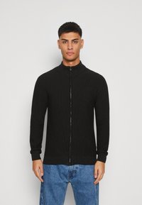 Svart glidelåsjakke med høy krage, med ribbet tekstur og lange ermer, paret med blå denimjeans.